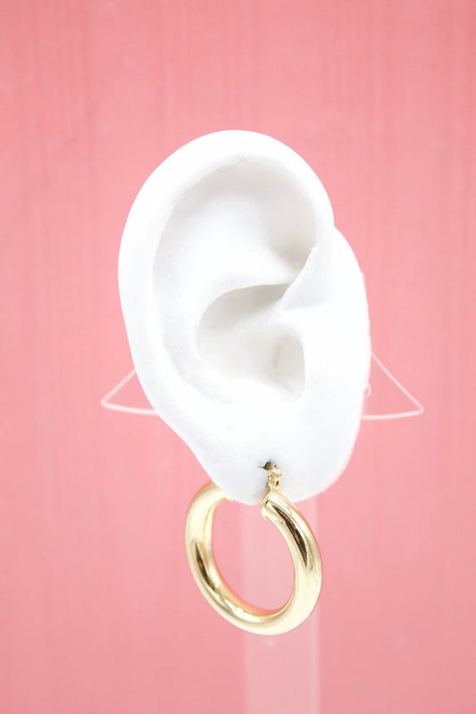 A white mannequin ear displays a thick gold hoop earring against a pink background.