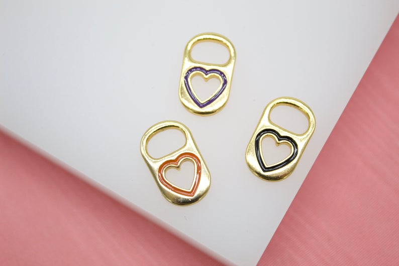 Three gold soda tab charms with colorful heart-cutout enamel details rest on a white surface against a pink background.