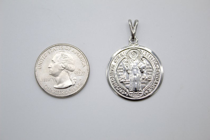 A silver Saint Benedict medal rests beside a US quarter on a plain white surface to show its relative size.