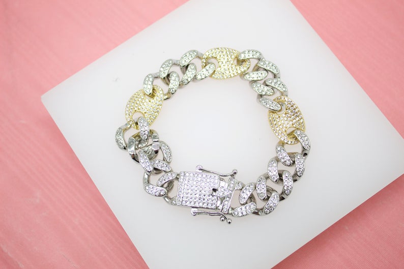 A sparkling cuban link bracelet featuring alternating silver-paved links and gold nugget accents rests on a white display surface against a pink background.