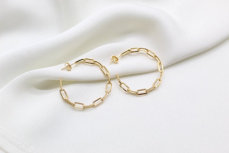 A pair of gold paperclip chain hoop earrings rests elegantly on a soft white silk background.