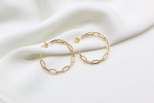 A pair of gold paperclip chain hoop earrings rests elegantly on a soft white silk background.