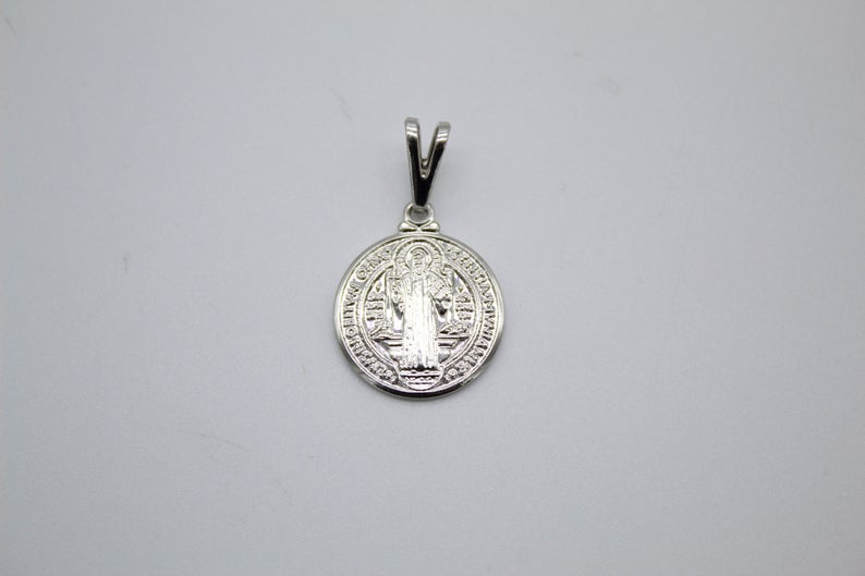 A polished silver Saint Benedict pendant rests flat on a plain white surface.