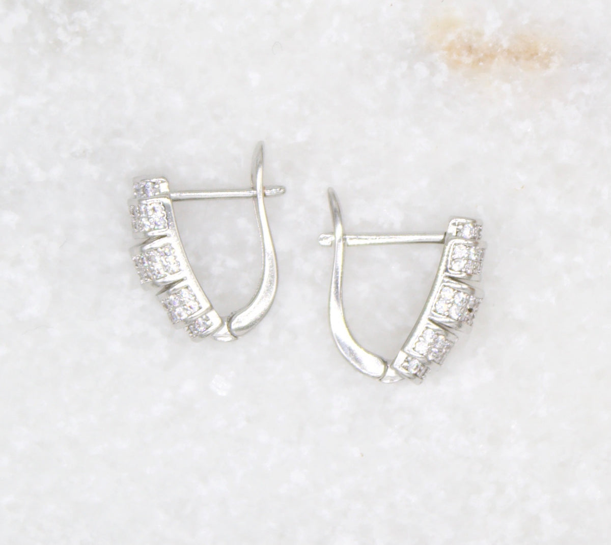A pair of silver huggie hoop earrings encrusted with small shimmering diamonds rests on a white marble surface.