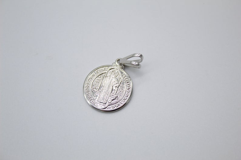 A silver Saint Benedict medal pendant rests on a neutral surface, showcasing intricate religious engraving on its circular face.