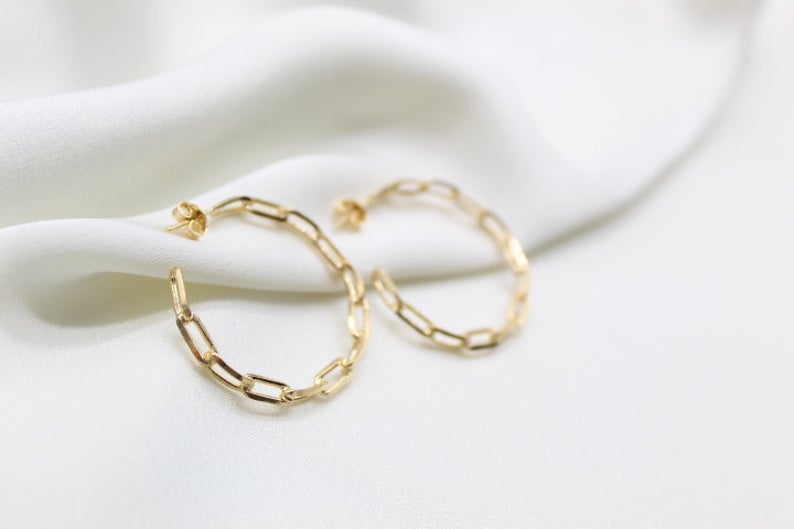 Two gleaming gold paperclip chain hoop earrings rest delicately on a soft white silk cloth.