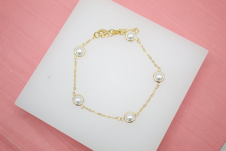 A delicate gold chain bracelet features bezel-set pearl charms resting on a white display surface.