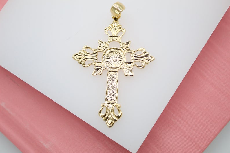 An ornate gold cross pendant rests on a white display surface, showcasing intricate filigree details and a textured center sunburst design.