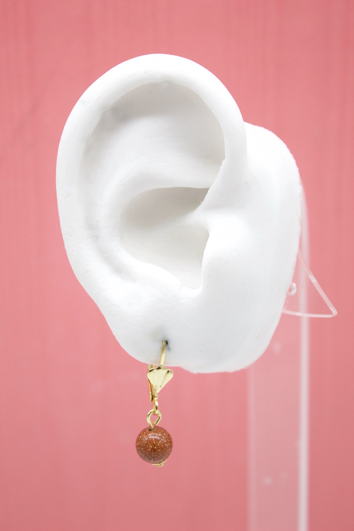 A white mannequin ear displays a gold leverback earring featuring a dangling, sparkling brown gemstone bead against a pink background.