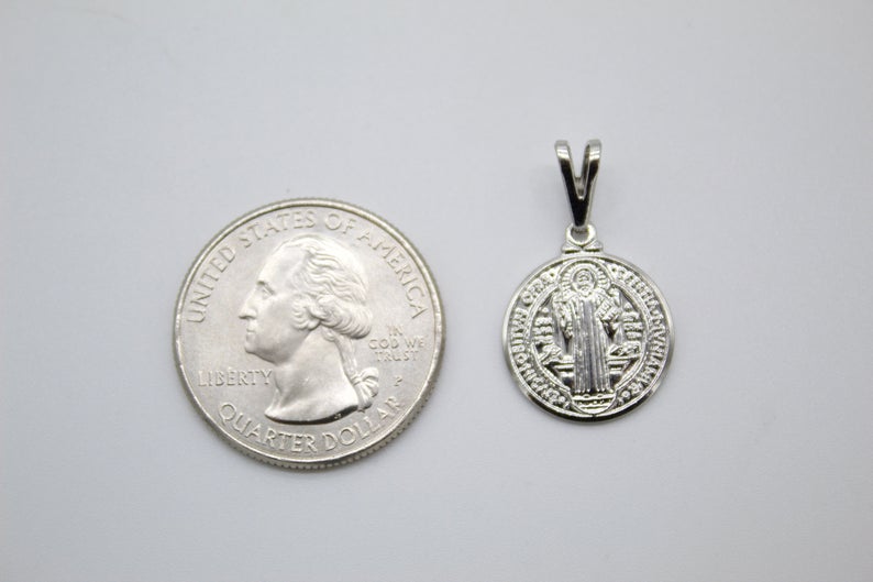 A sterling silver Saint Benedict medal pendant rests next to a US quarter coin to display its size.