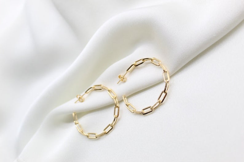 A pair of stylish gold chain-link hoop earrings rests elegantly on a soft white silk background.