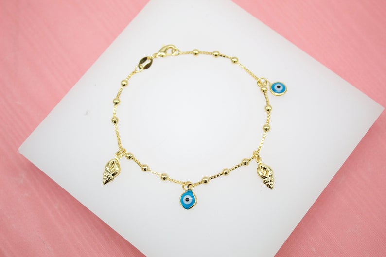 A delicate gold charm bracelet featuring alternating beads and dangling seashell and blue evil eye charms rests on a white display surface.