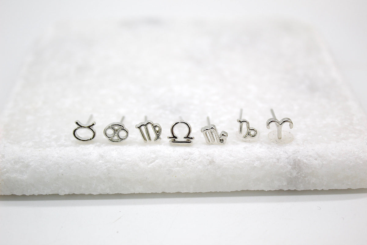 A collection of delicate silver zodiac sign stud earrings lines up neatly on a white marble surface.