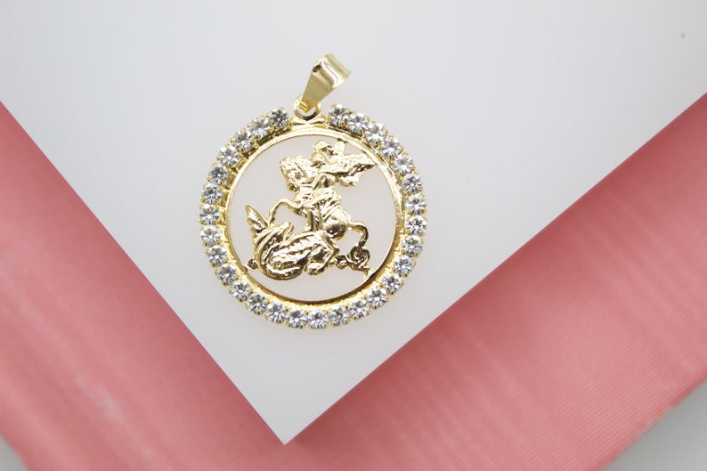 A round gold pendant featuring Saint George slaying the dragon rests on a white display surface, surrounded by a sparkling halo of crystals.