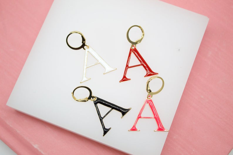 Four colorful letter A enamel charm hoop earrings rest on a white display surface against a pink background.