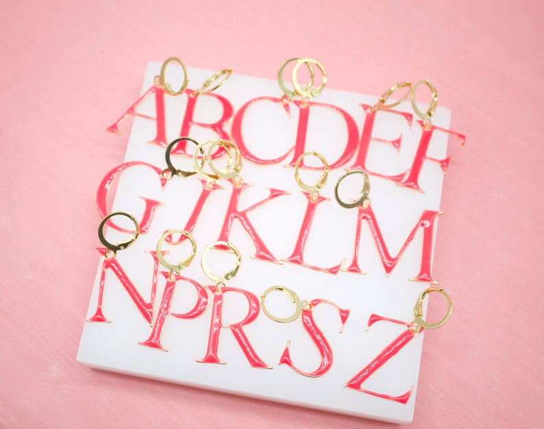 A collection of pink and gold enamel initial letter hoop earrings is neatly arranged on a white display surface against a soft pink background.