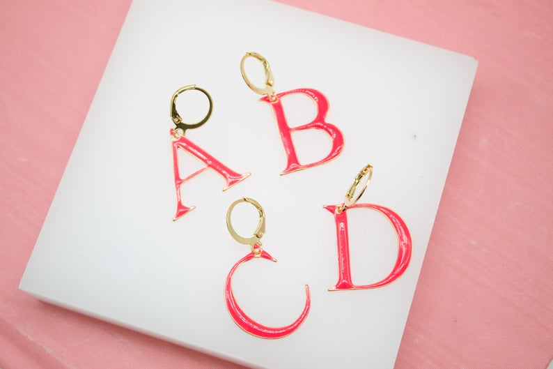 Four gold initial letter hoop earrings featuring vibrant pink enamel display the letters A, B, C, and D on a white display stand.
