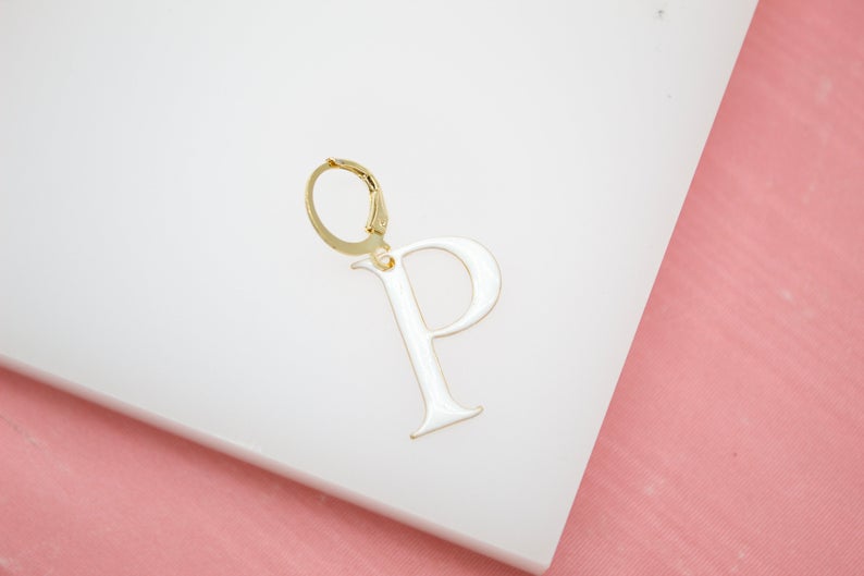 A gold initial P earring featuring a glossy white enamel letter rests against a white display surface.