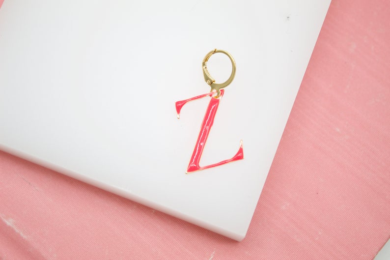 A gold initial Z earring featuring vibrant pink enamel rests on a white surface.