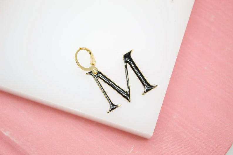 A stylish gold initial M hoop earring rests on a white surface next to a pink backdrop.