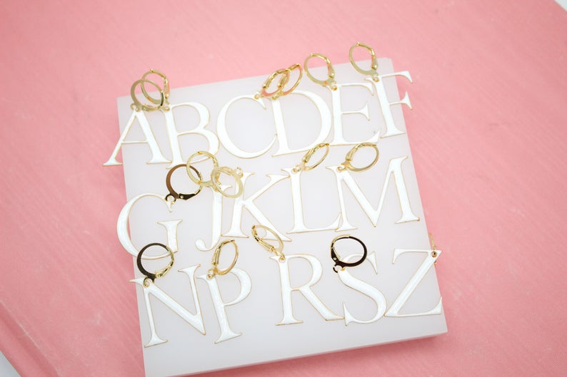 A collection of white acrylic alphabet letter earrings with gold hoop clasps rests on a white display board against a pink background.