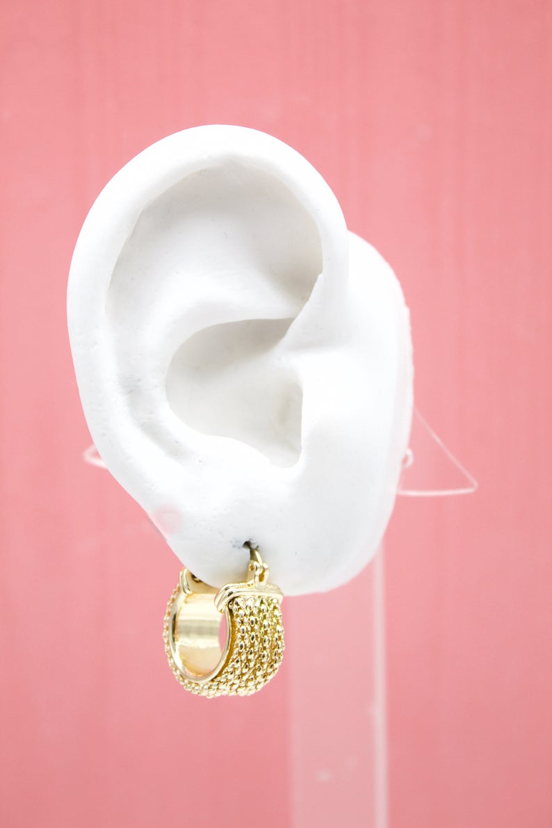 A white mannequin ear displays a textured gold hoop earring against a pink background.