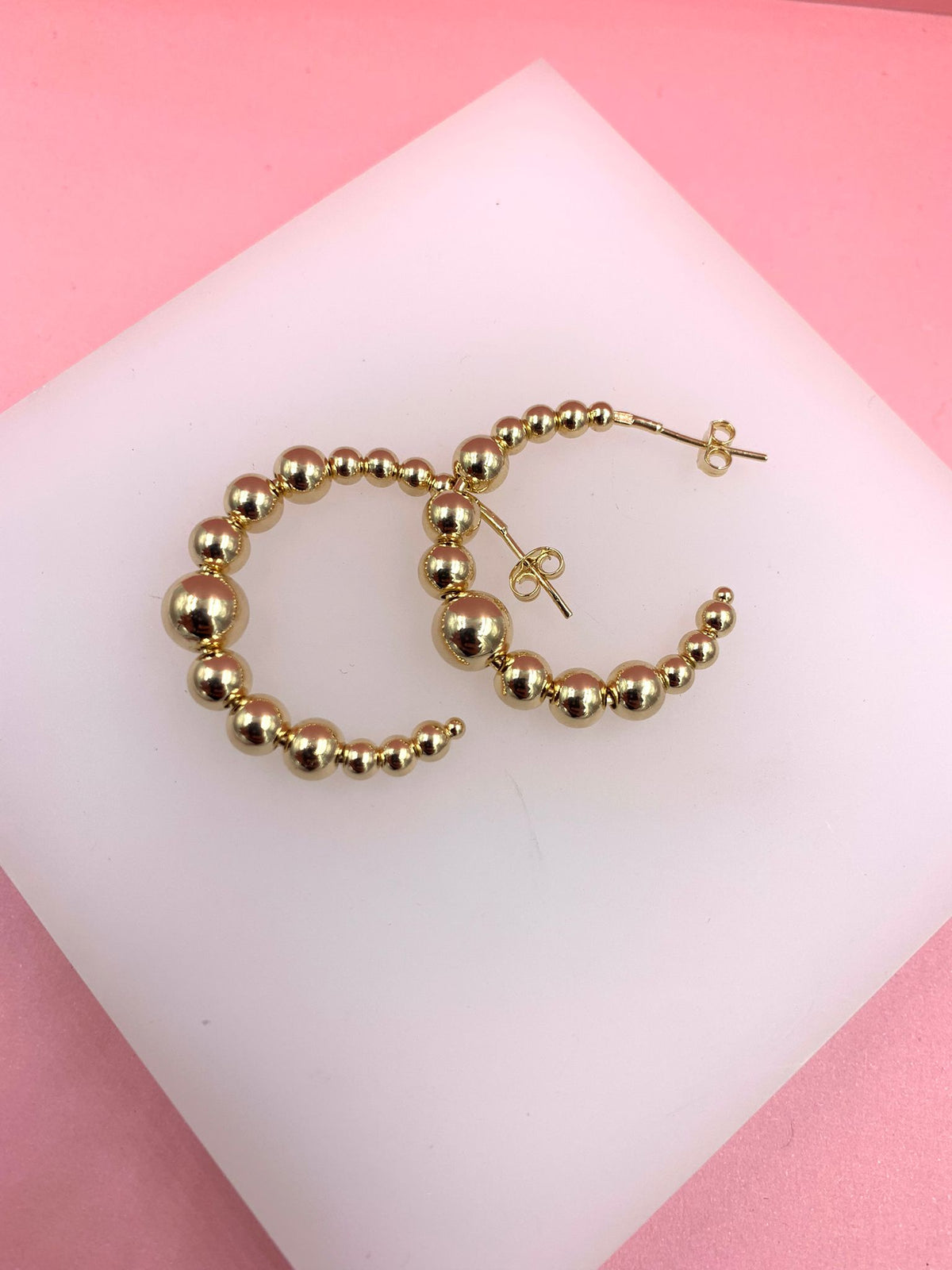 A pair of shiny gold ball hoop earrings rests on a white platform against a pink background.