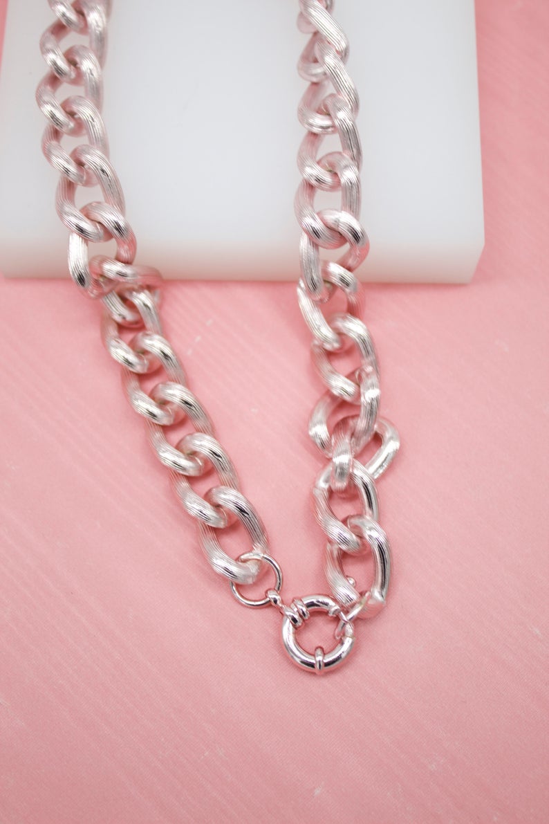 A textured silver chain necklace rests on a pink surface against a white backdrop.