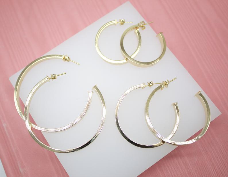 Three pairs of polished gold hoop earrings in varying sizes rest on a white display stand against a pink background.