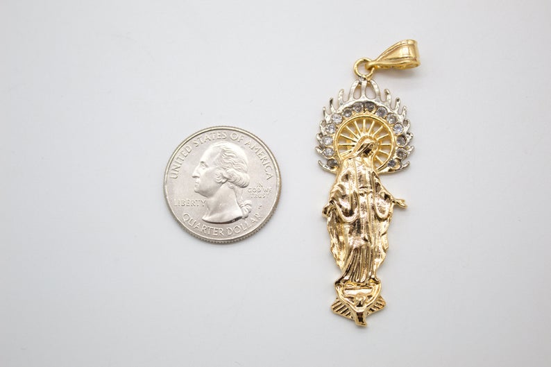 A gold and silver Virgin Mary pendant rests next to a US quarter coin for scale on a light surface.