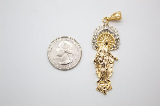 A gold and silver Virgin Mary pendant rests next to a US quarter coin for scale on a light surface.