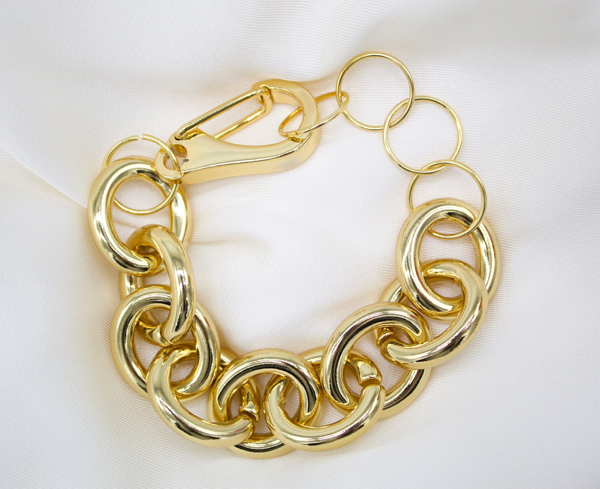 A chunky gold link bracelet rests on a white satin background, featuring large interlocking rings and a carabiner clasp.