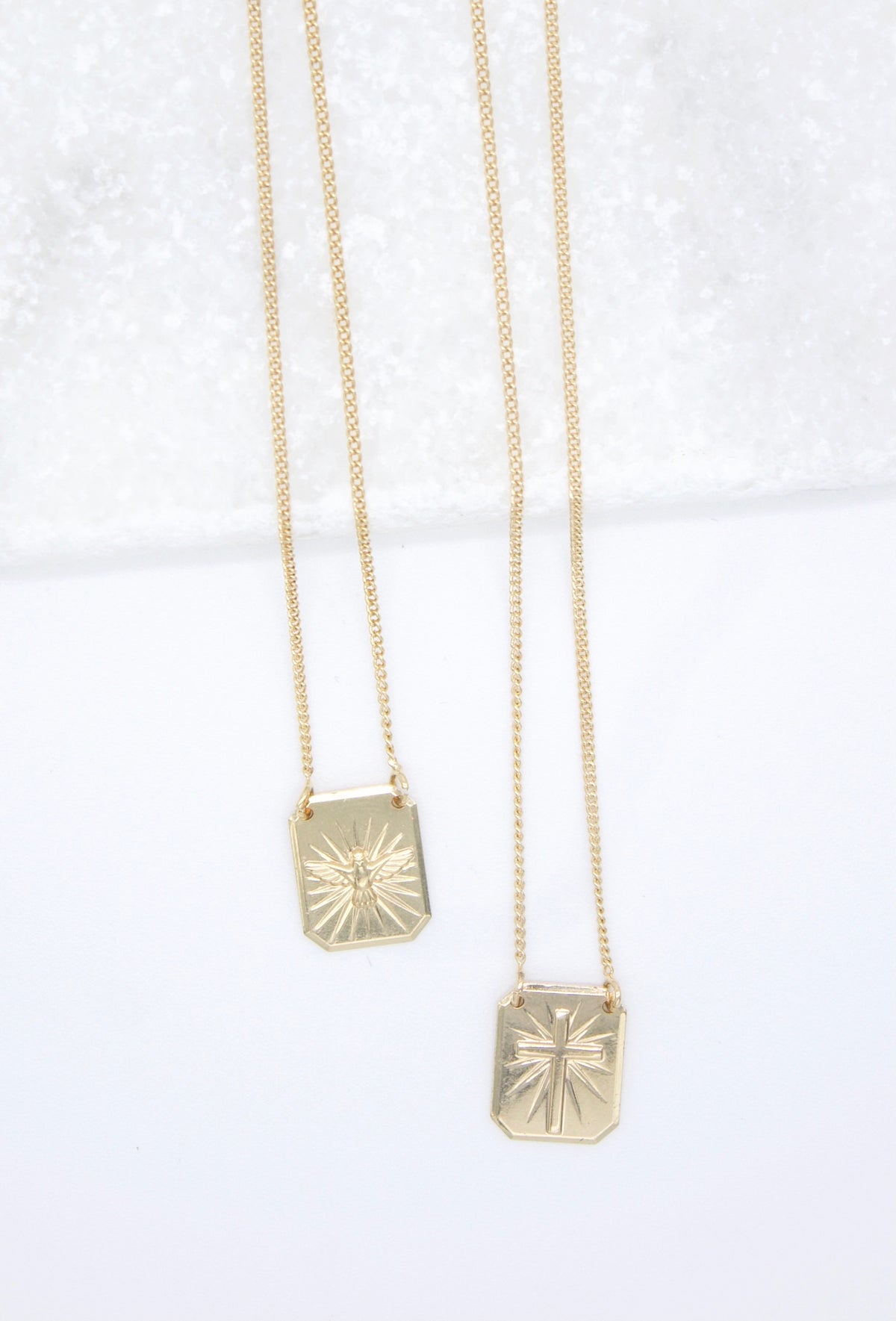Two delicate gold pendant necklaces featuring rectangular charms with embossed Christian symbols display against a white surface.