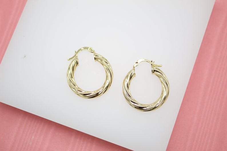 A pair of polished gold twist hoop earrings rests on a white display against a pink background.