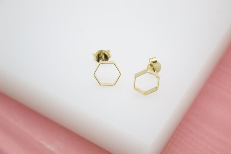 A pair of gold hexagon geometric stud earrings rests on a white display surface against a pink background.