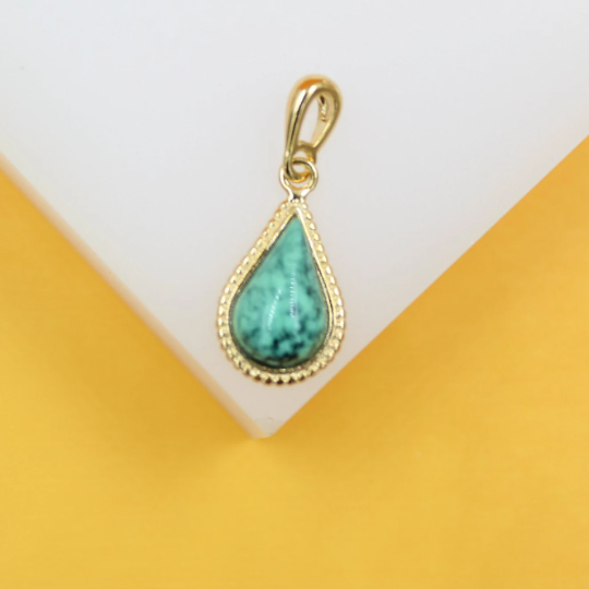 A teardrop-shaped turquoise gemstone pendant with a gold bezel setting rests on a white display against a vibrant yellow background.