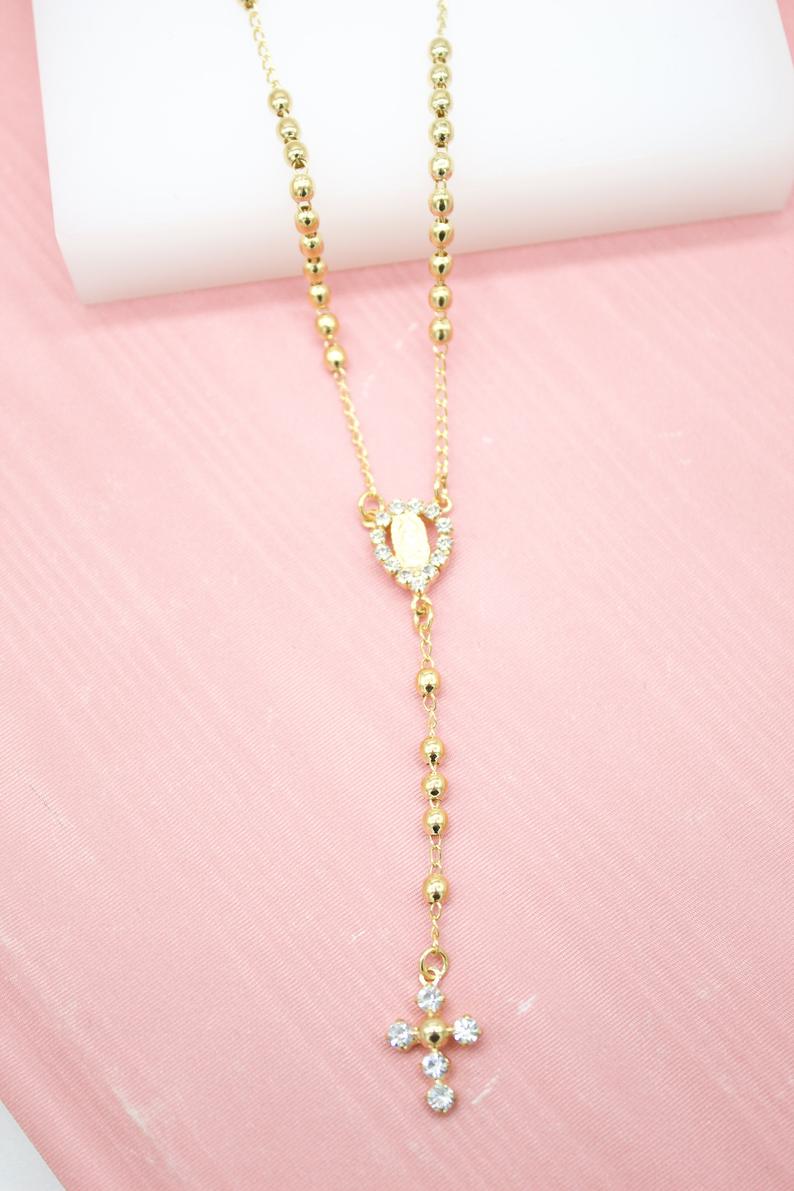 A delicate gold rosary necklace featuring bead spacers, a rhinestone-encrusted centerpiece, and a sparkling cross pendant rests on a pink surface.