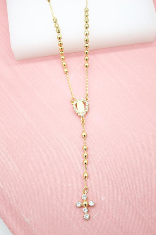 A delicate gold rosary necklace featuring bead spacers, a rhinestone-encrusted centerpiece, and a sparkling cross pendant rests on a pink surface.