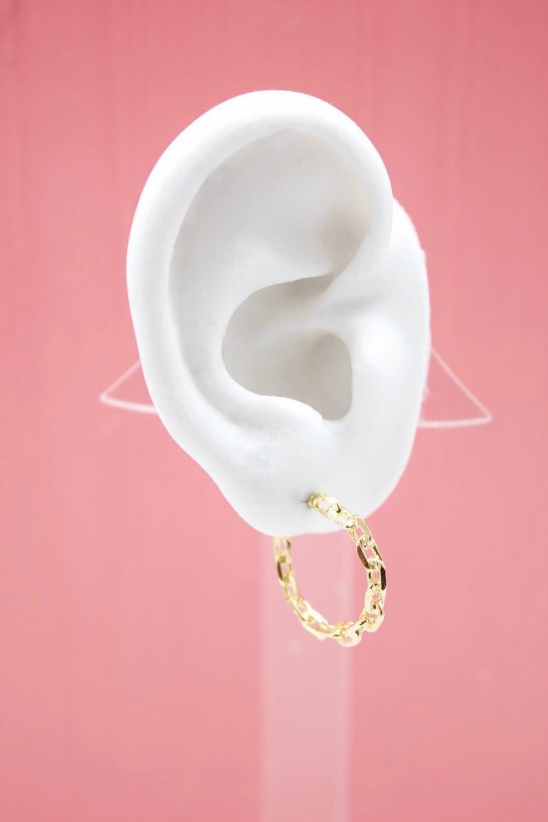 A white mannequin ear displays a shiny gold chain link hoop earring against a pink background.
