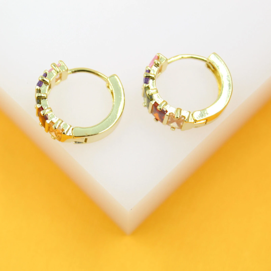 A pair of gold huggie hoop earrings featuring colorful multi-gemstones rest on a white display stand against a vibrant yellow background.
