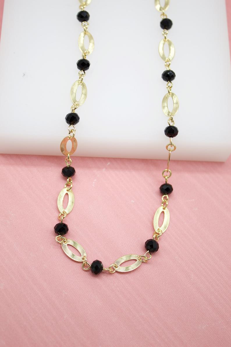 A chic gold-link necklace features alternating textured oval links and faceted black crystal beads against a soft pink and white background.