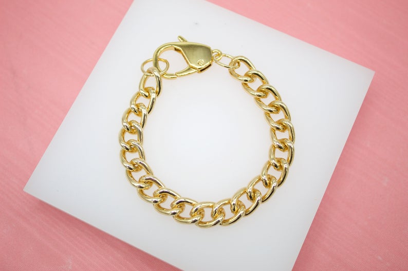 A gold curb chain bracelet with a lobster clasp rests on a white display surface against a pink background.