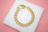 A gold curb chain bracelet with a lobster clasp rests on a white display surface against a pink background.