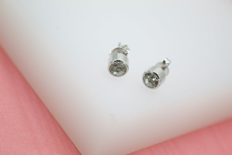 A pair of sparkling silver stud earrings rests on a white display block against a soft pink background.
