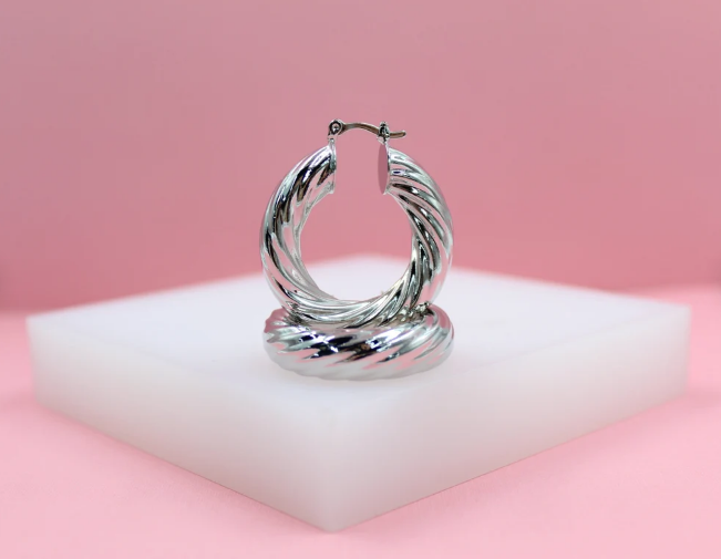 A pair of glossy silver twisted hoop earrings rests on a white display block against a soft pink backdrop.