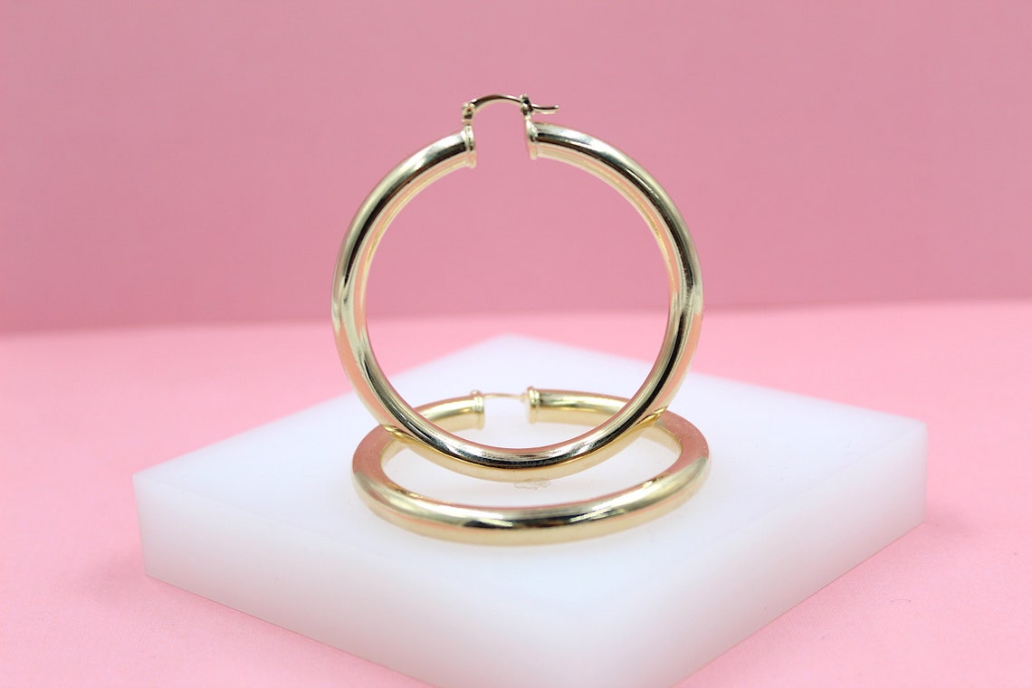 Two thick gold hoop earrings rest on a white display block against a pink background.