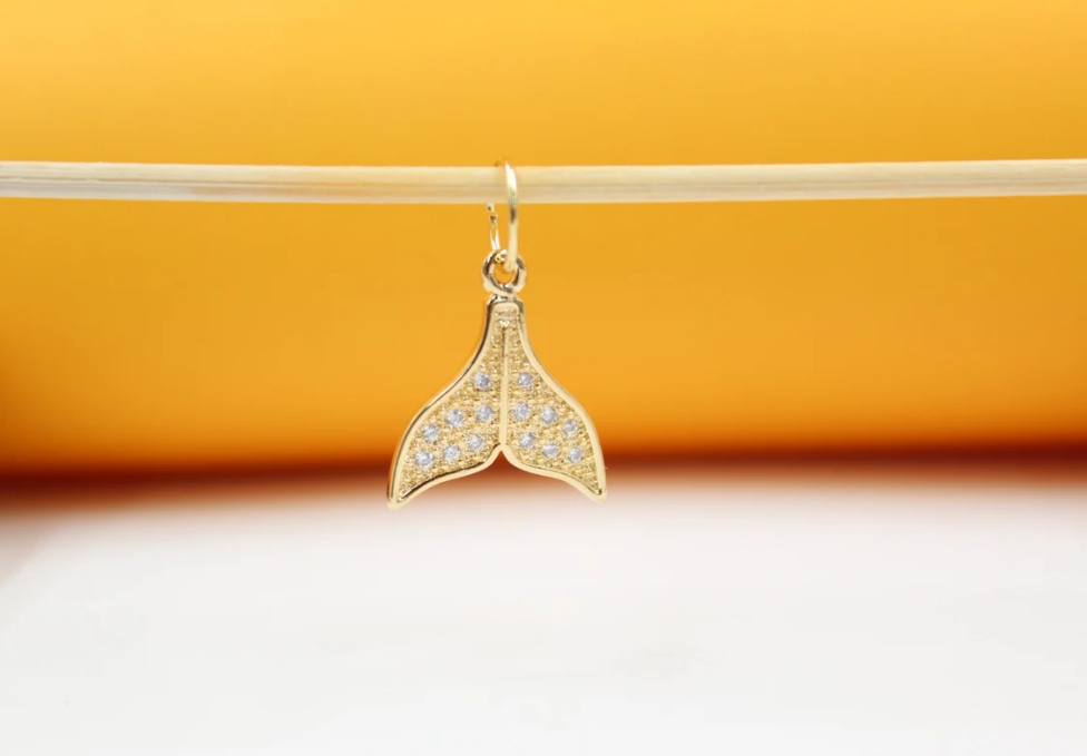 A gold whale tail pendant encrusted with sparkling cubic zirconia crystals hangs from a delicate stick against a warm yellow and white background.