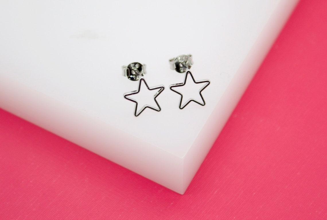 A pair of silver star-shaped stud earrings rests neatly on a white display block against a vibrant pink background.