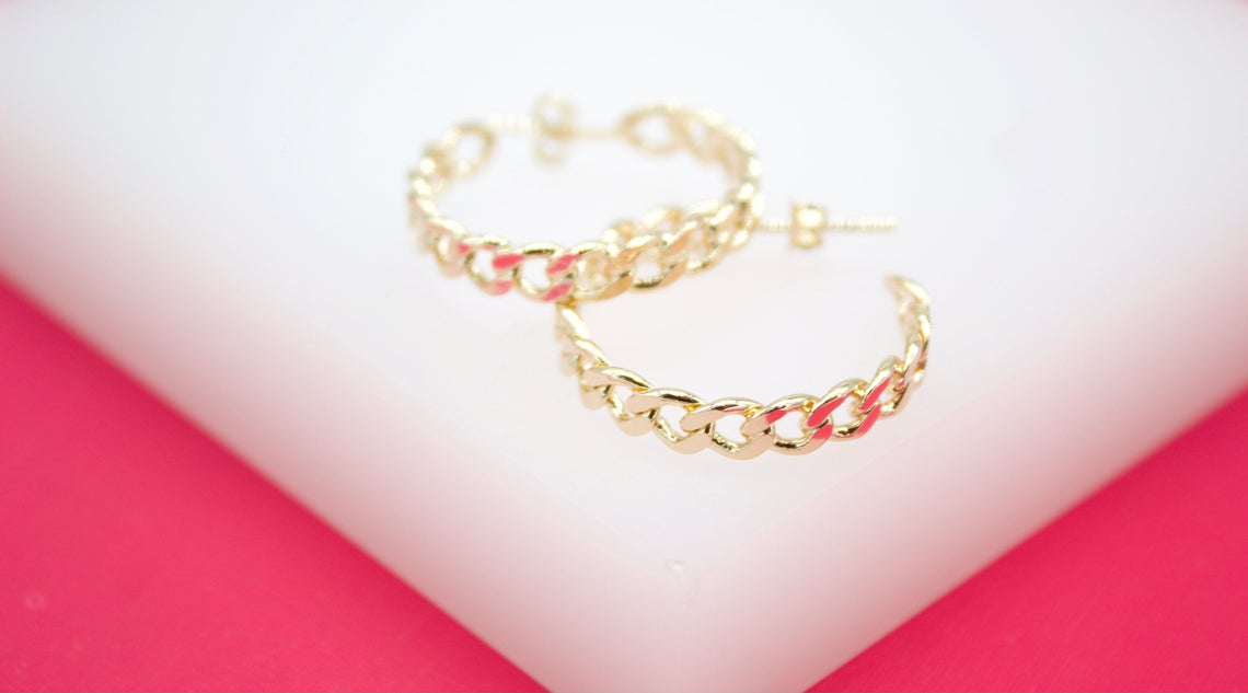 A pair of polished gold-plated cuban link hoop earrings rests on a white display surface against a vibrant pink background.