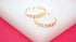 A pair of polished gold-plated cuban link hoop earrings rests on a white display surface against a vibrant pink background.